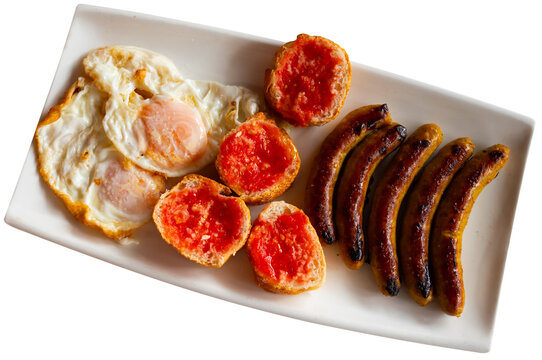 Mediterranean breakfast consisting of sunny side up eggs with spicy grilled butifarra sausages and traditional Spanish pan con tomato sandwiches is served on a white plate. Highlighted on a white