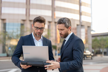 Two entrepreneurs near the office on the street. Business meeting of friends outdoors. Business mens with takeaway coffee and laptop outdoor. Business men team using laptop outdoor. Two businessman