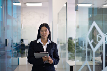 In a modern office hallway, a young businesswoman stands holding a tablet