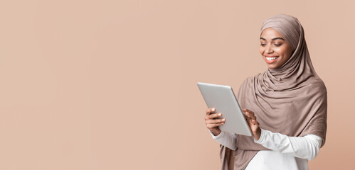 Portrait of smiling afro muslim girl in hijab using digital tablet, downloading new app or browsing media, standing over yellow background, free space