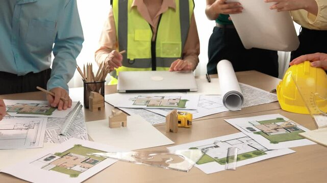 Smart architect and safety engineer discussion about building blueprint on table with architectural document, measurement tools and wooden block scatter around at modern office. Closeup. Delineation.