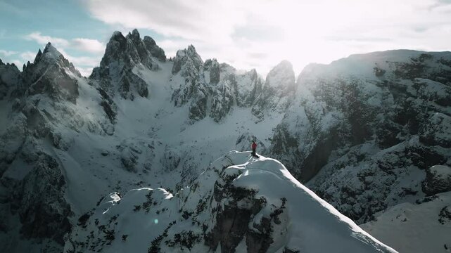 Aerial view of a snowy landscape with mountains in the background and with an hiker on top of a central viewpoint. A rare drone footage of Cadini di misurina viewpoint in winter, Dolomites, Italy