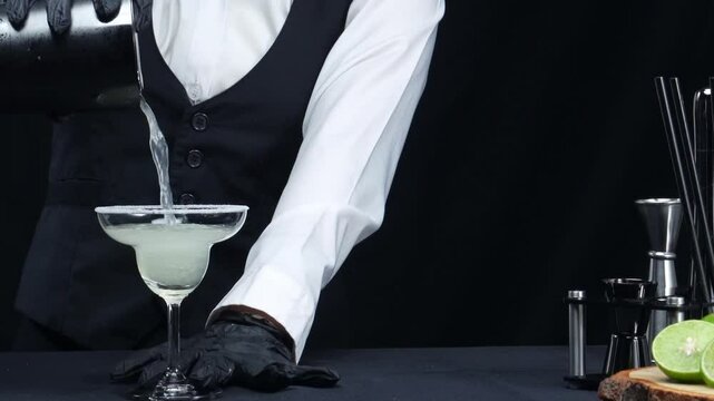 Professional bartender pouring fresh margarita with salted garnish glass. Macrography of skilled bartender hands prepare for making cocktail with black background at the bar. Slow motion. Comestible.