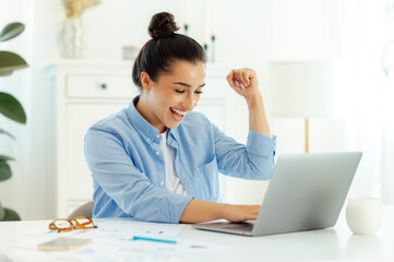 Joyful happy caucasian young woman, company seo rejoice in victory while sitting at the work desk with laptop, gesturing with her hands, rejoicing in a good deal, big profit, great results, smile