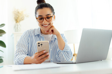 Pretty, happy busy young caucasian woman with glasses, sitting on a workplace with a laptop, uses...