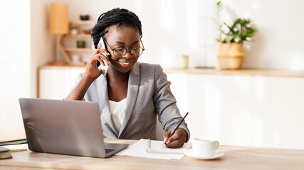 Black Millennial Businesswoman Talking On Cellphone And Taking Notes While Working On Laptop In...