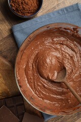 Chocolate dough in bowl, spoon and cocoa on wooden table, flat lay