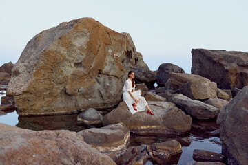serene woman in flowing white dress relaxes on rocky outcrop in water, red shoes peeking out