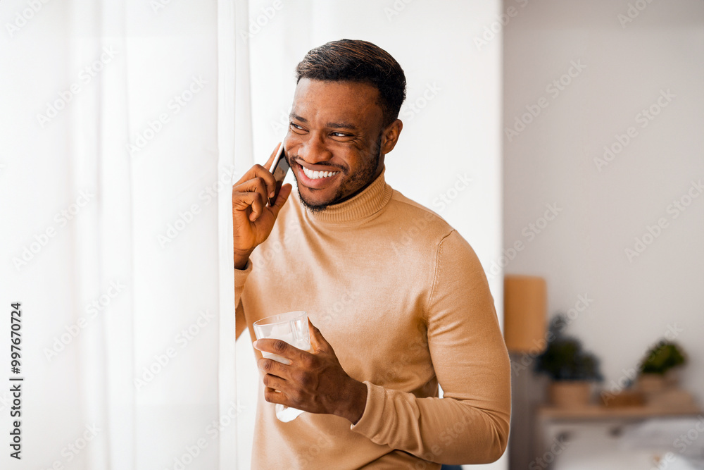 Poster phone call. joyful afro guy talking on cellphone having coffee standing next to window at home. copy