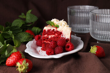 Piece of red velvet cake on plate, glass, berries and mint on brown background