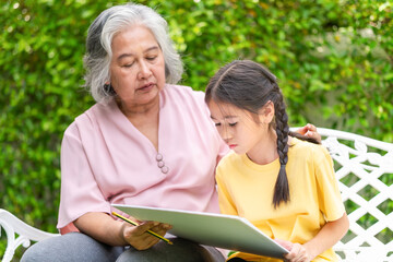 Senior Asian woman and her granddaughter play together in the garden, Grandmother with kid girl drawing picture in the yard