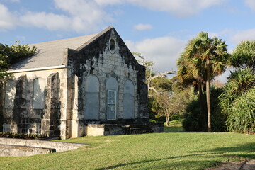 This photograph captures a historic stone building surrounded by lush greenery, bathed in warm, late-afternoon sunlight. The weathered structure features arched windows and a metal door, showing signs