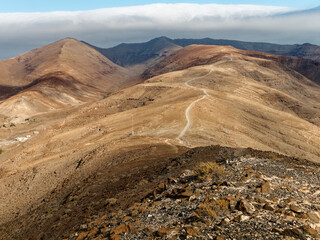 The trail to the summit of Pico de la Zarza, the highest mountain on Fuerteventura, winds through arid, rocky terrain. The undulating landscape reveals distant hills, with a cloud-topped horizon in th
