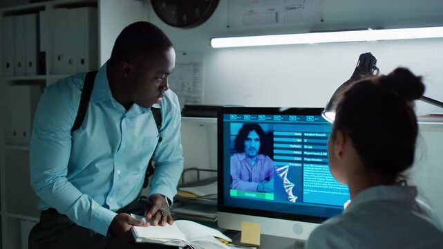 African American male investigator pointing at computer screen and discussing suspect profile with female forensic scientist while working together in laboratory