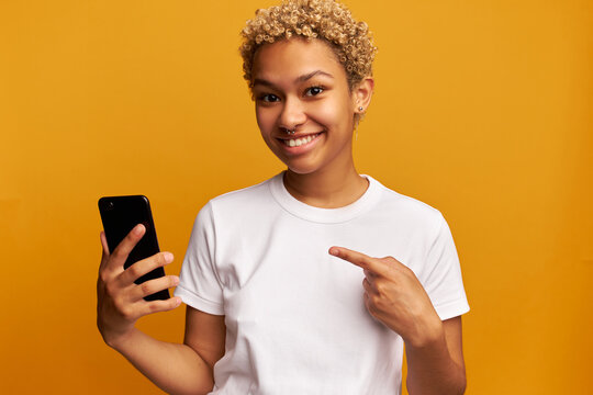 Young african american stylish female promoting new application or mobile game, pointing at smartphone in her hands, recommending you to download and try online study services, isolated on yellow