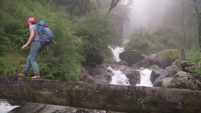 Woman with backpack and trekking poles crossing mountain river wooden log bridge during Makalu Barun National Park trek in Nepal. Mountain hiking and active people concept image.