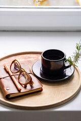 Cozy workspace with notebook, glasses and coffee cup on wooden tray.