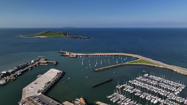 Howth Harbour, County Dublin, Ireland, May 2023. Drone pushes forward above the harbour Marina over the Pier and Lighthouse with Ireland's Eye in background with Lambay Island in the distance.