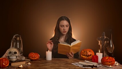 Halloween woman witch fortune teller sits at table reads from notebook spell on candle, being possessed by spirit, wind blows off flame. Magic halloween concept.