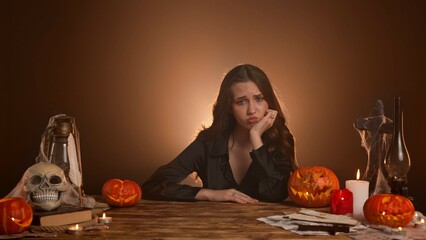 Halloween gothic woman witch fortune teller sitting at table looks around, bored tired expression, yawning sleepy face. Magic halloween concept.