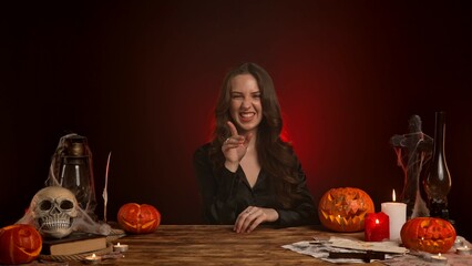 Halloween gothic woman witch fortune teller sits at table looks around playfully smiling, shows quiet silence gesture finger over mouth. Magic halloween concept.