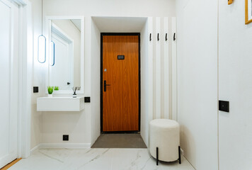 In a spacious hallway, a beautifully crafted wooden door leads to another room, a reflective mirror is on the wall, and a stylish white ottoman is neatly placed on the floor