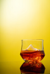 Glass of liquor scotch whiskey with lemon and ice cubes on bright yellow background and reflective table top. Space for text