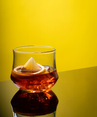Glass of liquor scotch whiskey with lemon and ice cubes on bright yellow background and reflective table top. Space for text