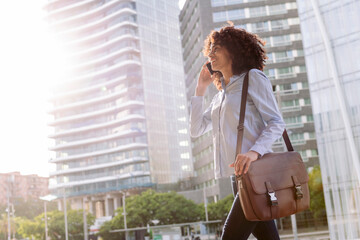 smiling ethnic female entrepreneur walking in downtown and talking on mobile phone while looking away