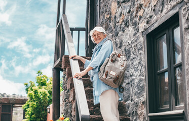 Smiling traveler senior woman in outdoor excursion in rustic mountain village looking at camera. Elderly female enjoys healthy lifestyle and activity