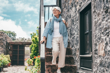 Fototapeta premium Smiling traveler senior woman in outdoor excursion in rustic mountain village looking away. Elderly female enjoys healthy lifestyle and activity