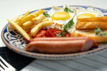 Breakfast plate with fried eggs, sausage, tomatoes, and fries served on a decorative blue plate
