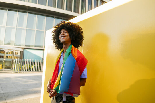 Thoughtful gay man with LGBTQIA flag day dreaming near yellow wall