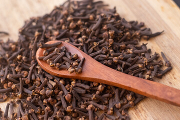 A close-up image of a pile of dried cloves on a wooden spoon. The cloves are a dark brown color and have a distinct, knobby shape. They are clustered together, showing their intricate textures