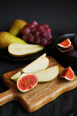 Ripe whole and sliced ​​figs, pears and pink grapes on a wooden board and black plate on a dark background