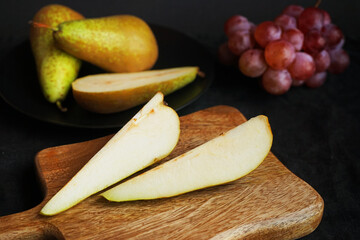 Ripe whole and sliced ​​pears on a wooden board and plate next to pink grapes on a dark background