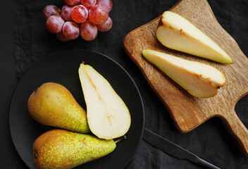Ripe whole and sliced ​​pears on a wooden board and plate next to pink grapes on a dark background