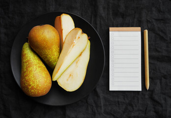 Ripe sliced ​​and whole pears on a black plate next to a blank to-do list on a dark background