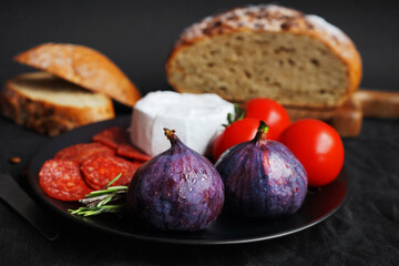 Plate with appetizers of Brie cheese, perroni, tomatoes, figs and rosemary sprigs next to sliced ​​grain bread on a dark background