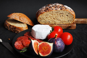 Plate with appetizers of Brie cheese, perroni, tomatoes, figs and rosemary sprigs next to sliced ​​grain bread on a dark background