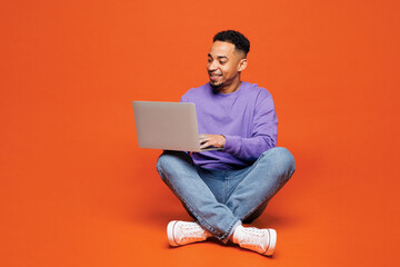Full body young happy IT man of African American ethnicity wear purple sweatshirt casual clothes hold use work on laptop pc computer isolated on plain red orange background studio. Lifestyle concept.