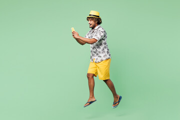 Full body side view young cool Indian man wear beach shirt hat rest near hotel pool jump high hold use mobile cell phone isolated on plain green color background. Summer vacation sea sun tan concept.