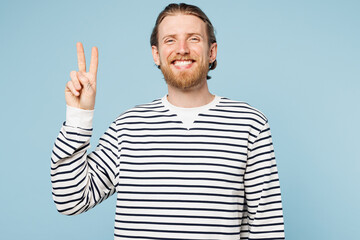 Naklejka premium Young smiling cheerful cool happy man he wear striped sweatshirt casual clothes showing victory sign look camera isolated on plain pastel light blue cyan background studio portrait. Lifestyle concept.