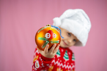 A cheerful toddler in a Christmas sweater and Santa hat holds an ornament on a pink background, conveying holiday joy and excitement with his smile. Christmas, New Year.