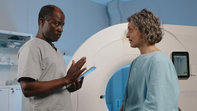 Black radiographer holding digital tablet working with mature Caucasian female patient in MRI room