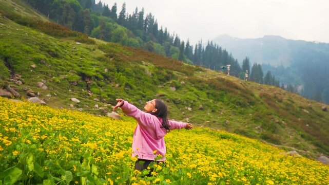 Indian teenager girl standing in between yellow flowers on mountains at Manali, Himachal Pradesh, India. Girl raising hands and enjoying nature. Blooming wildflowers during spring in Himalayas