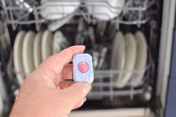 A woman holds a capsule of detergent against the backdrop of a dishwasher with dirty dishes.