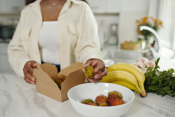 Person holding open box of fresh rambutan on kitchen counter with bananas and greenery in background, preparing for a healthy meal