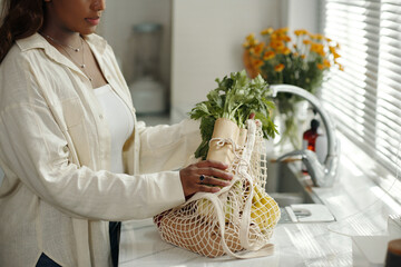 African American woman holding a reusable mesh bag with fresh vegetables standing in bright kitchen with sunlit surroundings