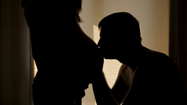 Silhouette of couple against background of window in house. Husband lovingly kisses belly timmy of his pregnant wife. Waiting for miracle and birth of baby. He strokes and puts his ear to his stomach